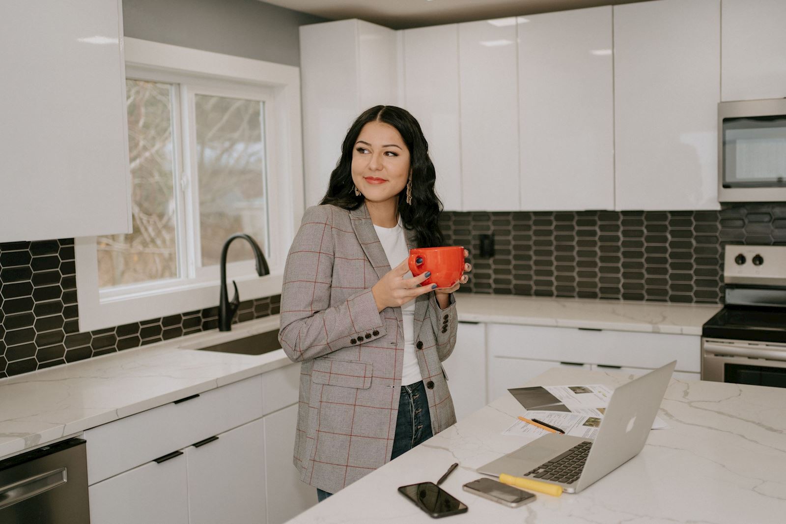Sarah Aguirre standing in a bright kitchen with a coffee cup, laptop, and home documents.